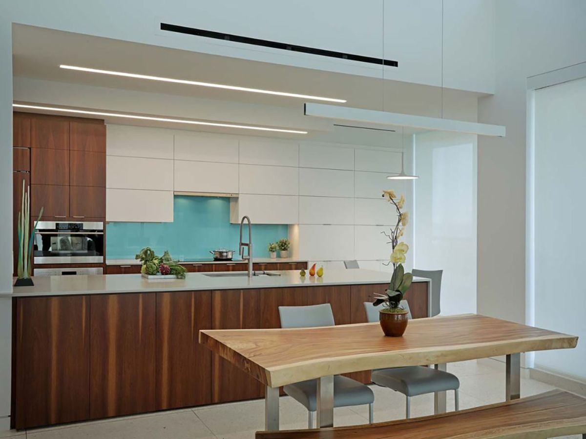 Modern Sarasota kitchen with custom two-tone cabinetry (walnut-look and white flat panels), vibrant teal glass backsplash, white waterfall island, live-edge wood table, and contemporary linear lighting, exemplifying high-end design by a premier Sarasota cabinetry designer.